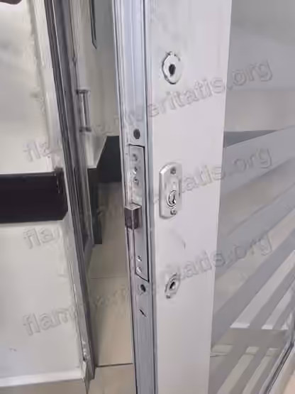 Canto de una puerta corrediza de vidrio y aluminio en el aula 2107, edificio 2, con una chapa de seguridad y dos orificios de montaje vacíos, sin manija instalada.